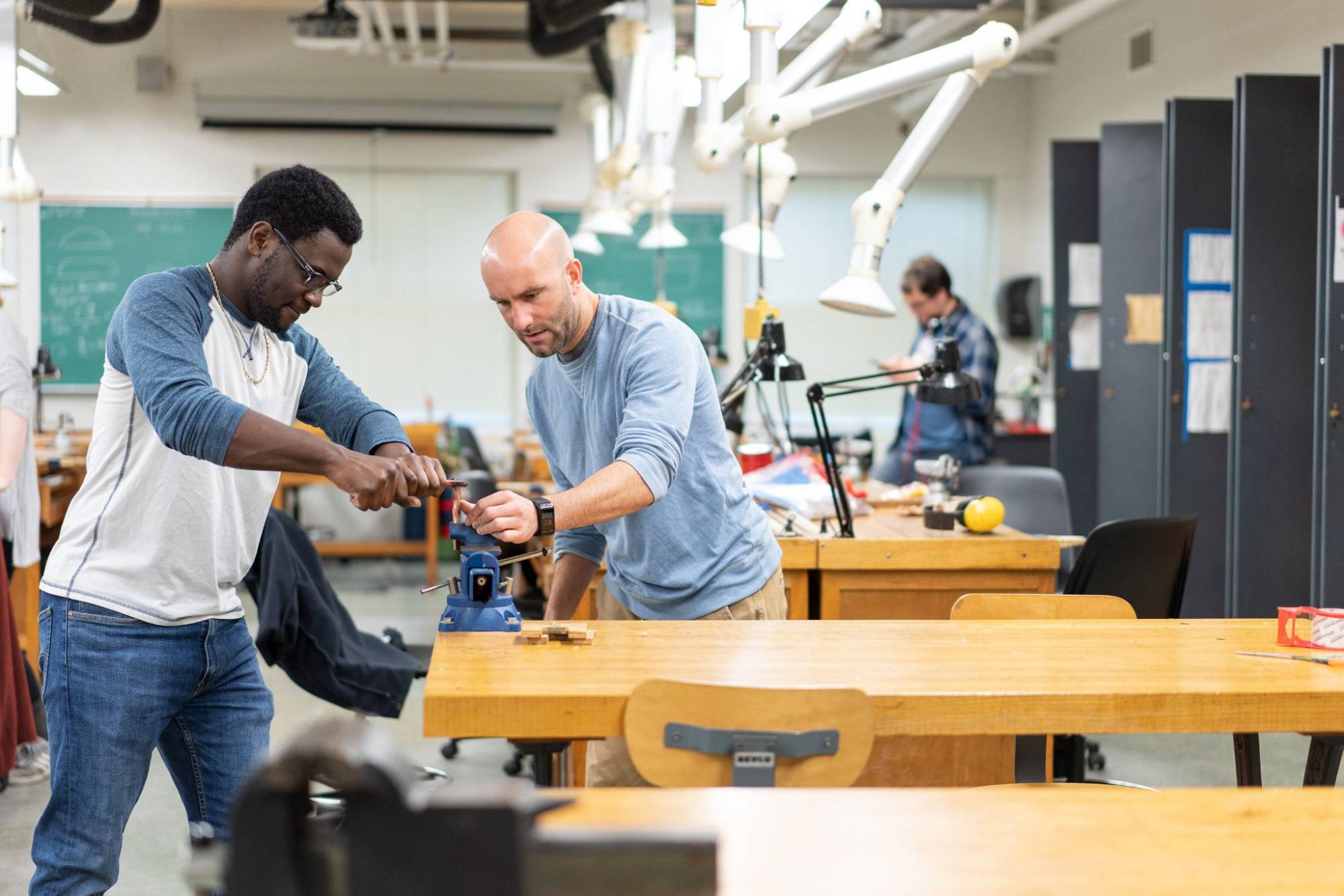 Image of a Black male student using equipment with the professor, Peter Antor, in a jewelry-making workshop. In the background there is more equipment and a young white male student.